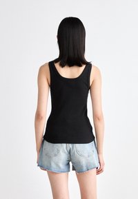 Kvinna med rakt svart hår iförd en svart ärmlös topp och ljusblå denimshorts, vänd bort mot en helt vit bakgrund.