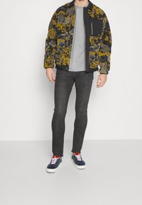 Wielobarwna, wzorzysta kurtka puchowa z kieszenią na zamek, szara koszulka, ciemne dżinsy i wielobarwne trampki.