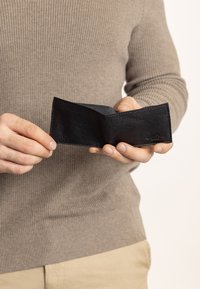 Schwarze Lederbörse, die in einer Hand gehalten wird, mit einer strukturierten Oberfläche und einem einfachen Falt-Design. Keine sichtbaren Beschläge oder Muster.