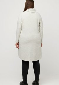 Vestido suéter de manga larga en gris claro con cuello vuelto, textura acanalada y dobladillo curvado, combinado con pantalones ajustados negros y botas de tobillo.
