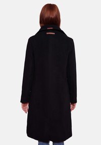 Una persona con lunghi capelli rossi indossa un cappotto nero di media lunghezza, in piedi con le spalle rivolte alla fotocamera su uno sfondo chiaro e uniforme.