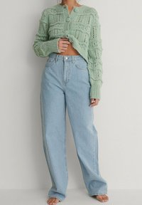 Mintgrön stickad cardigan med texturerade mönster, knäppning framtill, tillsammans med ljusblå höga jeans. Barfota mot en neutral bakgrund.