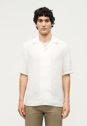 Jeune personne aux cheveux bouclés foncés portant une chemise blanche à manches courtes et un pantalon brun clair, debout devant un fond uni clair.