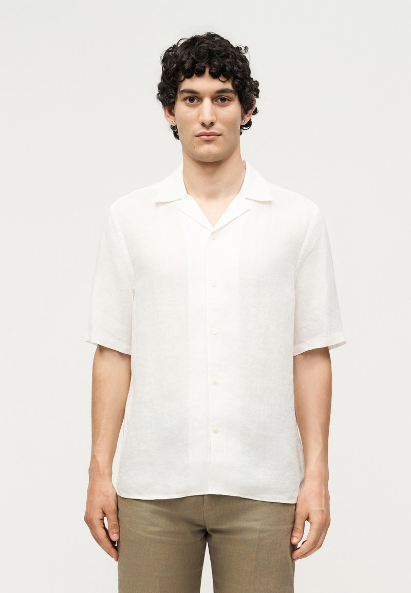 Νεαρό άτομο με σγουρά σκούρα μαλλιά που φοράει λευκό πουκάμισο με κοντά μανίκια και ανοιχτό καφέ παντελόνι, στέκεται μπροστά σε απλό φωτεινό φόντο.