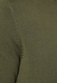 Un tejido de punto en verde oliva presenta una textura uniforme con un sutil patrón acanalado y un detalle de costura visible a lo largo de un borde.