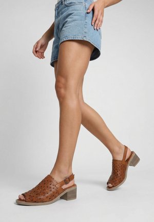 Femme portant un short en denim bleu et des sandales en cuir marron à talons perforées, marchant sur un fond gris clair.