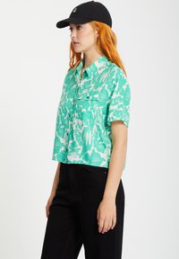 Kortsleeve, kort skjorta i grön och vit abstrakt mönster, med knäppning fram och två bröstfickor, kombinerad med svarta byxor.