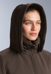 Hnedý kapucňový kabát so zipsom vpredu, s jemným kožušinovým golierom a hladkým, matným povrchom. Detailný pohľad na tvár.