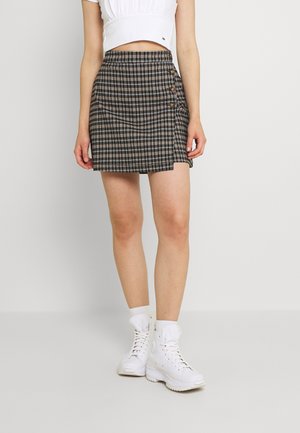 Kvinde iført en brun ternet miniskørt med høj talje og sidelommer med knapper, hvid crop top, hvide sokker og hvide høje sneakers, stående.