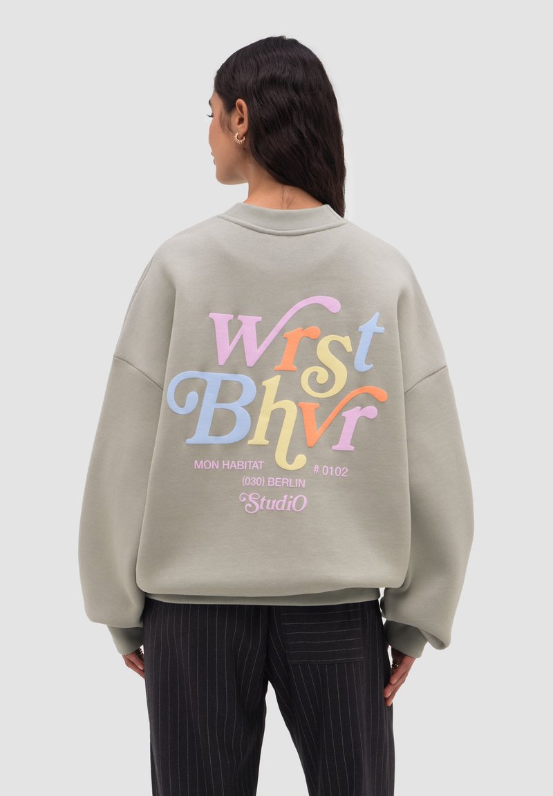 Mujer con cabello largo y oscuro que lleva una sudadera beige de gran tamaño con texto estilizado y colorido y pantalones negros de rayas finas, de pie frente a un fondo gris.