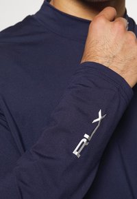 En mans handled och hand som justerar en mörkblå långärmad skjorta med en silverfärgad "RLX"-logotyp på ärmen.