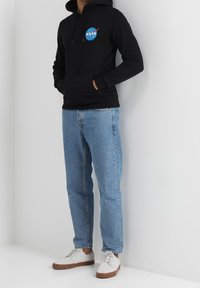 Sudadera con capucha negra con el logo de la NASA, jeans de mezclilla azul claro y zapatillas blancas con suelas marrones, de pie contra una pared blanca.