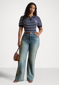 Sieviete ar gariem cirtainiem matiem, ģērbusies tumši zilu svītrainu polo krekliņā, plašā bikšu modelī ar izbalējušu krāsu, brūnā jostā, sandales kājās, un rokā tur nelielu brūnu somiņu.