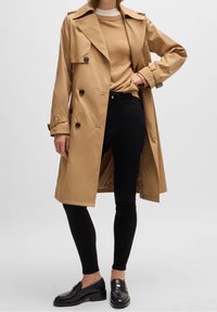 Kobieta ubrana w beżowy płaszcz trenchowy, pod którym ma sweter w odcieniu kamelowym, czarne wąskie spodnie i czarne mokasyny, stoi z jedną ręką na biodrze.