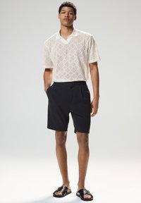 Fiatal férfi áll egy egyszerű szürke háttér előtt, bézs mintás rövid ujjú pólót, fekete rövidnadrágot és fekete papucsot viselve.