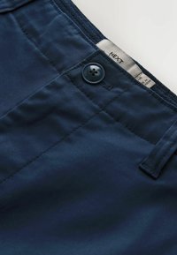 Prim-plan al taliei pantalonilor bleumarin, cu un nasture cusut și o etichetă de material vizibilă în interior, pe care scrie „NEXT.”