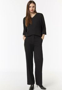 Black loose-fitting top with V-neck, paired with high-waisted black trousers. Both in smooth fabric, model wears black heeled ankle boots.