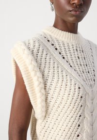 Svetr z pleteniny v off-white odstínu, s kabelkovými akcenty na rameni a texturovaným, otevřeným designem s žebrovaným límcem.
