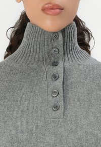 Gros plan sur une personne portant un pull à col roulé en ribbed gris avec six boutons foncés sur le devant.