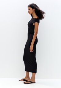 Schwarzes, knielanges Kleid mit Raffung an den Schultern, aus weichem Stretchmaterial, figurbetont geschnitten, kombiniert mit schwarzen flachen Sandalen. Minimalistisches Design.