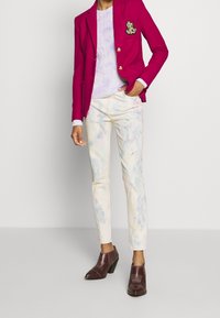 Fuksjowa marynarka z złotym emblematem, lawendowy sweter z grubym warkoczem, pastelowe dżinsy z plamami oraz brązowe botki na obcasie z teksturowanymi akcentami.