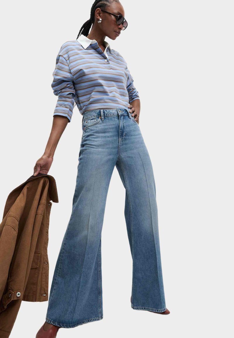 Jasnoniebieskie szerokie dżinsy o prostym kroju, w zestawie z wielokolorową, długą bluzką z rękawami z biało-niebieskimi paskami i białym kołnierzykiem, noszoną z brązową kurtką.