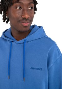Sudadera azul con capucha, que presenta un tejido texturizado, cordones azules contrastantes y un logo bordado en el pecho.