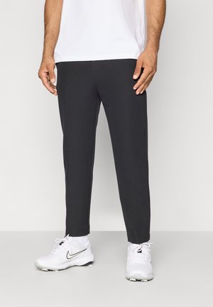 Man wearing black tapered pants, white socks, and white Nike sneakers standing against a plain light background.