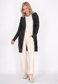 Tmavě šedý dlouhý cardigan s texturovaným povrchem, nošený přes bílý top a krémové kalhoty s širokými nohavicemi, doplněný o béžové kotníkové boty.