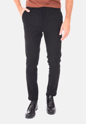 Uomo che indossa pantaloni in velluto a coste neri slim-fit con tasche laterali e scarpe stringate in pelle nera, in piedi davanti a uno sfondo bianco.