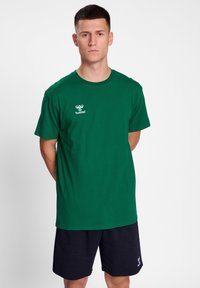 Groene katoenen t-shirt met ronde hals en korte mouwen, met een wit logo op de linkerborst. Draagt met donkere shorts.