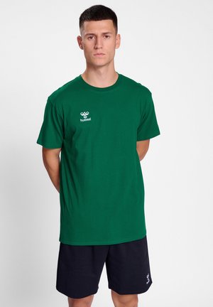 Groene katoenen t-shirt met ronde hals en korte mouwen, met een wit logo op de linkerborst. Draagt met donkere shorts.