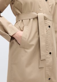 Μπεζ παλτό τύπου trench με ζώνη, διαθέτει τσέπη, ελαφρώς γυαλιστερή υφή και μαύρα κουμπιά κατά μήκος του μπροστινού μέρους.