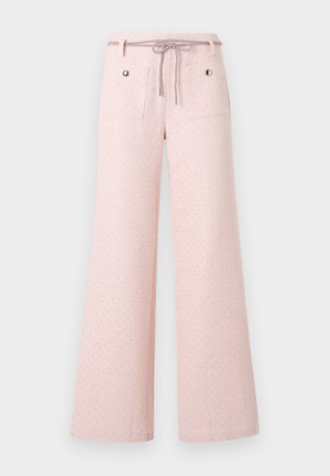 Žena v bielom tielku bez rukávov, svetloružových voľných nohaviciach s prednými vreckami a bielych nízkych špicatých topánkach, stojaca pred bielym pozadím.