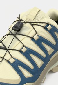 Hellgelber Sportschuh mit blauen Akzenten, strukturiertem Material und schwarzen elastischen Schnürsenkeln. Verfügt über eine geformte Sohle und einen sicheren Schnürmechanismus.