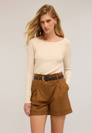 Vrouw met lang blond haar draagt een crèmekleurig, ribbelig longsleeve top, bruine high-waisted shorts en een donkere riem met een gouden gesp.