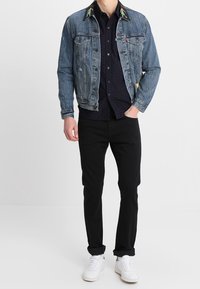 Ljusblå denimjacka med broderade krage detaljer, svart mönstrad skjorta under, svarta byxor och vita sneakers.