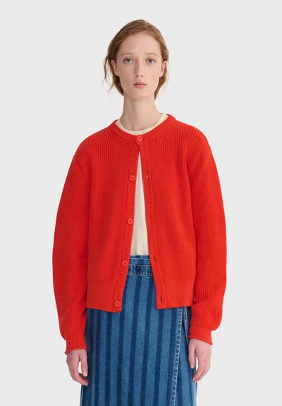 Femme portant un cardigan en maille rouge vif boutonné sur un haut blanc et une jupe en denim rayée bleue, debout devant un fond uni.