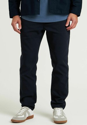Marineblaue Baumwollhose mit schmalem Schnitt, ausgestattet mit einer glatten Textur und klassischem Design, kombiniert mit hellgrauen und weißen Sneakers.