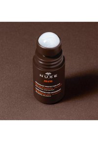 Contenant de déodorant cylindrique marron avec un capuchon roll-on blanc. Présente un texte orange indiquant la marque et les détails du produit. Texture de surface lisse.
