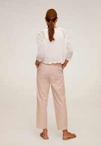 Kobieta z kucykiem, ubrana w biały sweter dzianinowy, beżowe spodnie z wysokim stanem i brązowe sandały, stoi z rękami w kieszeniach, odwrócona plecami.