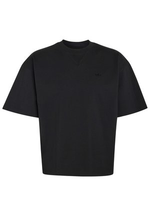 Ung mand iført sort oversized T-shirt, sort baglæns kasket og beige bukser, står mod en simpel lys baggrund.