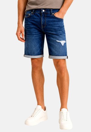 Mann trägt dunkelblaue hochgekrempelte Jeans-Shorts mit kleinem Rissdetail am rechten Oberschenkel und weiße Sneakers. Eine Hand in der Tasche.