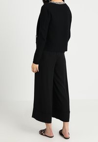 Czarny sweter o fakturowanym wykończeniu, z kontrastującym, wzorzystym kołnierzem. W zestawie z czarnymi spodniami o szerokich nogawkach, widziany od tyłu.