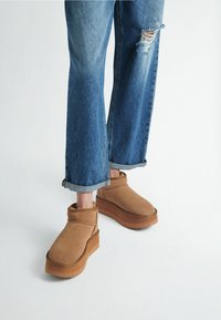 Brown suede ankle boots with a flat, thick rubber sole, paired with distressed blue denim jeans rolled at the cuffs.