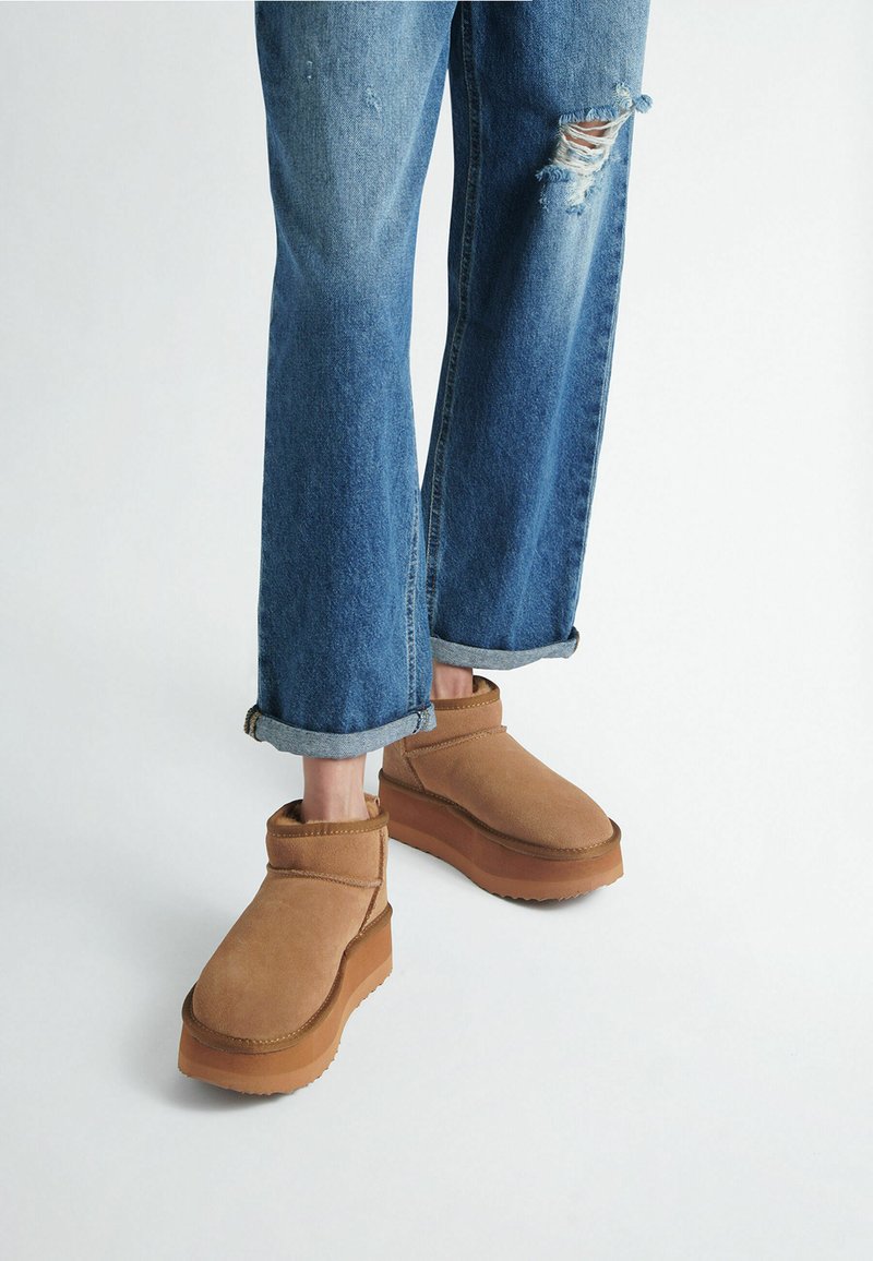 Brown suede ankle boots with a flat, thick rubber sole, paired with distressed blue denim jeans rolled at the cuffs.