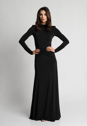 Kobieta w długiej, czarnej dopasowanej sukience z marszczeniami w talii i przy rękawach, stojąca z rękami na biodrach na tle jednolitego szarego tła.