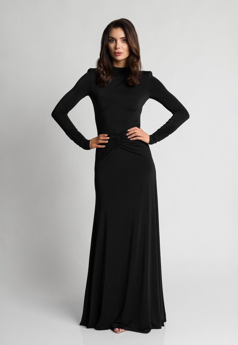 Kobieta w długiej, czarnej dopasowanej sukience z marszczeniami w talii i przy rękawach, stojąca z rękami na biodrach na tle jednolitego szarego tła.