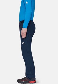 Granatowe spodnie outdoorowe wykonane z elastycznego materiału, z zamykanymi na zamek bocznymi kieszeniami, zaprojektowane z myślą o swobodzie ruchów i komforcie, w połączeniu z czarnymi i czerwonymi butami.