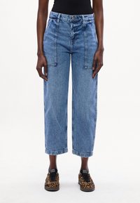 Jeans de mezclilla azul de pierna ancha con cintura alta, bolsillos frontales y costuras de contraste; combinados con zapatos negros y de estampado de leopardo.